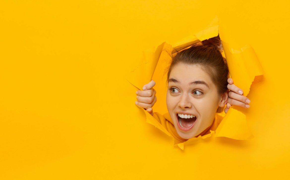 Excited girl looking through hole in paper at left with eyes round with surprise, isolated on yellow background with copy space