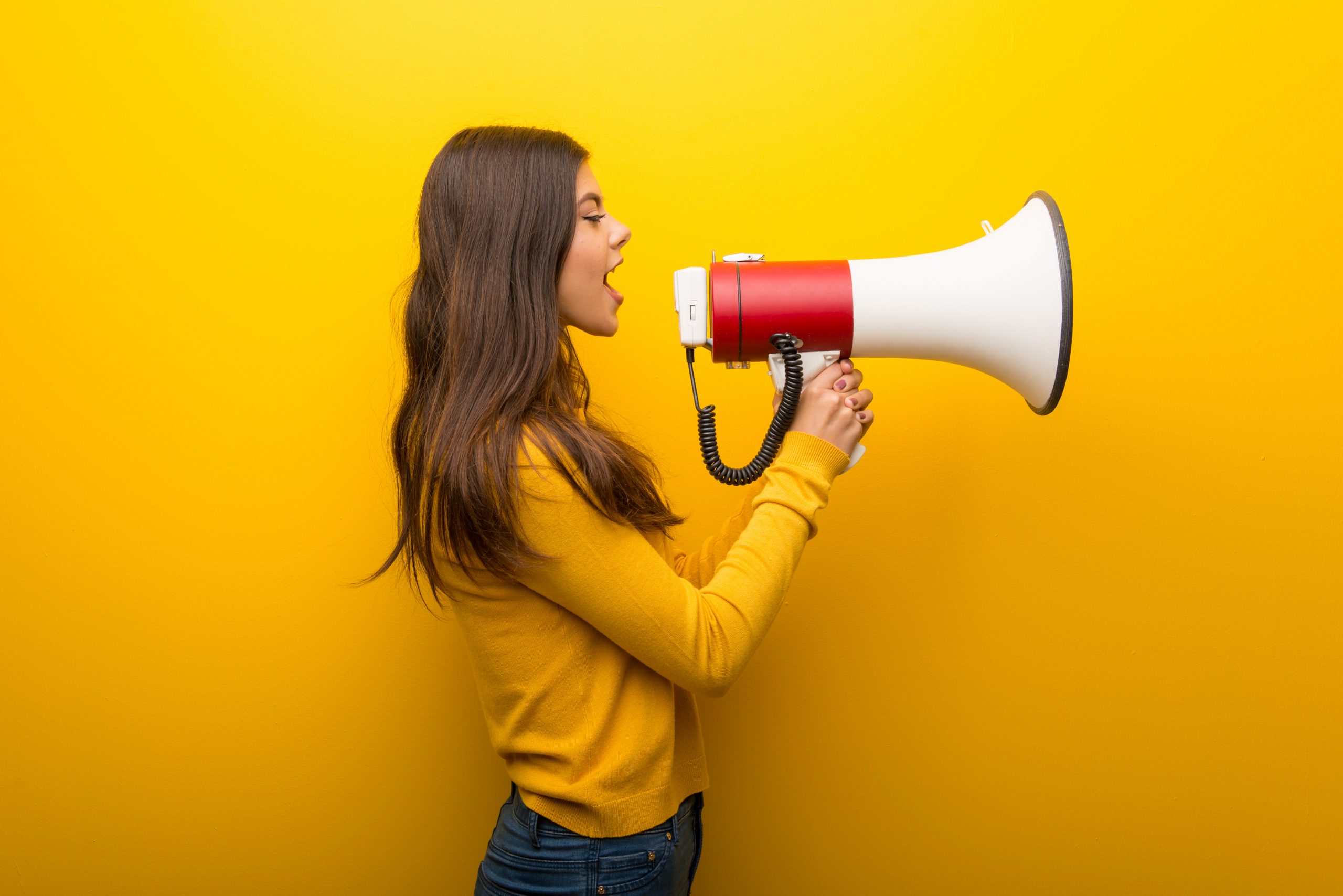Teenager girl on vibrant yellow background shouting through a megaphone to announce something in lateral position